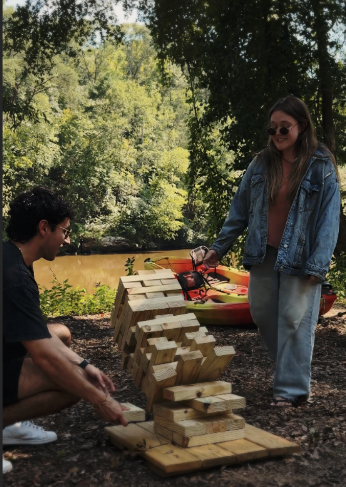 Two people outdoors near a river, building a large wooden Jenga tower with blocks. The woman is standing, wearing sunglasses, a denim jacket, and loose pants, smiling. The man is crouched, wearing glasses and a black t-shirt, focused on the game. A k
