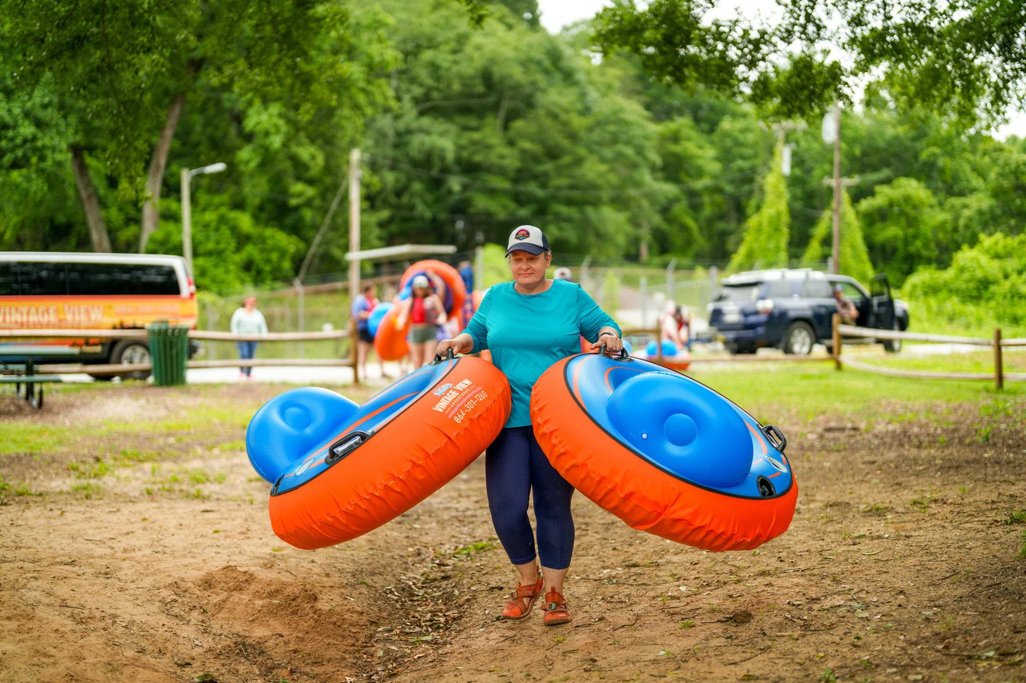 A woman holding two rescue tubes walking on grass at a summer outdoor event with green trees, a parked van, and other people in the background.
