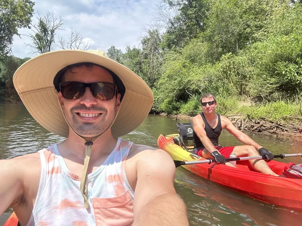 Two men kayaking on a river surrounded by green trees, one taking a selfie and the other paddling in a red kayak, both wearing sunglasses and summer clothes.
