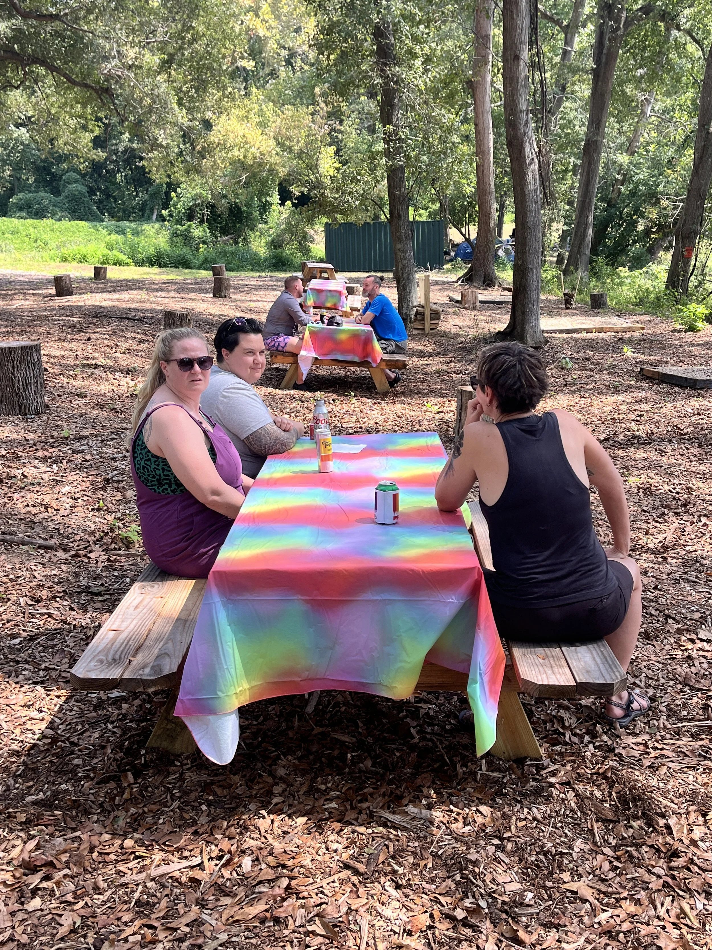 Four people sitting at picnic tables with rainbow-colored tablecloths in a wooded outdoor area, with trees and leaves on the ground.