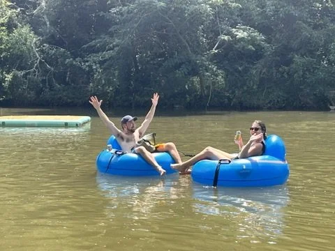 Two people relaxing on blue inner tubes in a lake, one waving and the other sipping a drink, with greenery in the background.