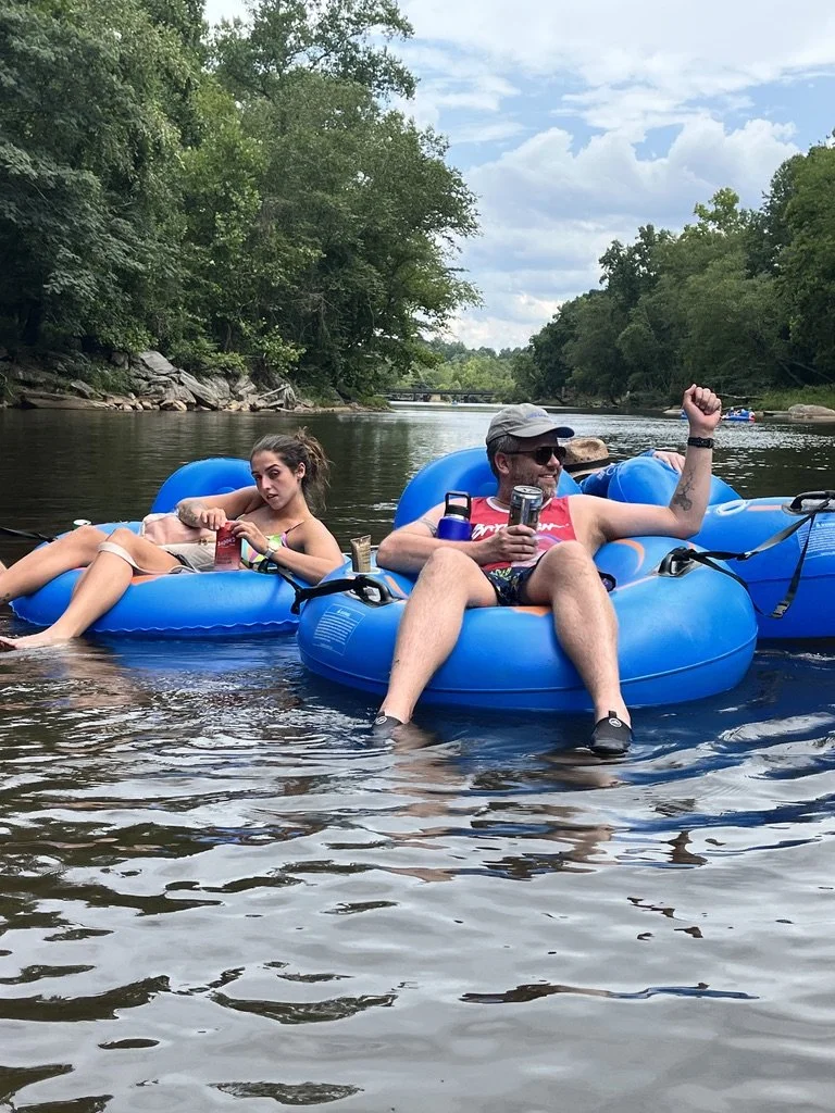 Two people relaxing on blue inflatable tubes floating on a river, surrounded by trees and a partly cloudy sky, holding drinks and enjoying a sunny day.