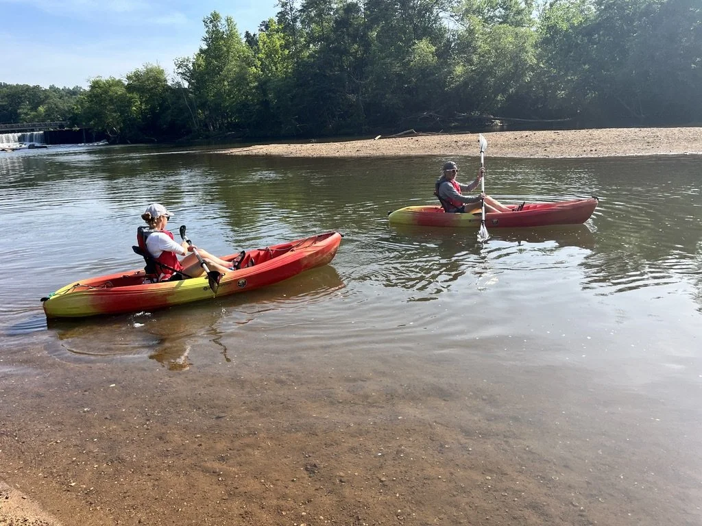 Two people kayaking on a calm river with a sandy shoreline, green trees, and a waterfall in the background on a sunny day.