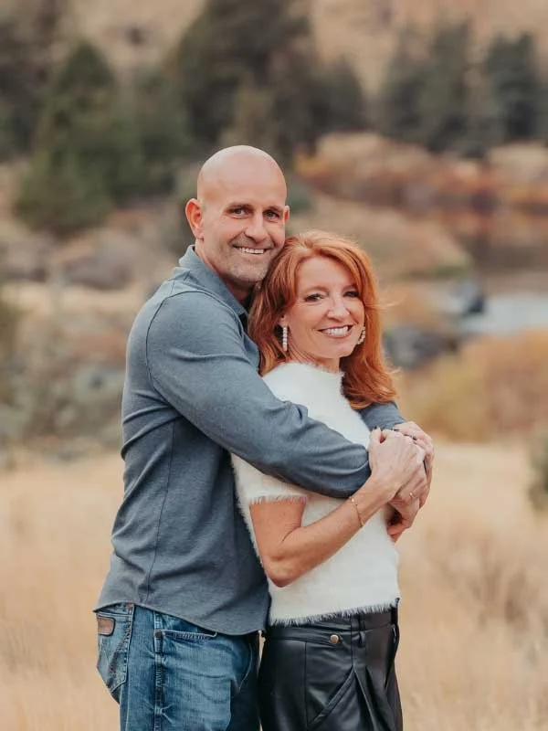 A smiling couple embracing outdoors in a natural setting with rocky terrain and sparse vegetation in the background.