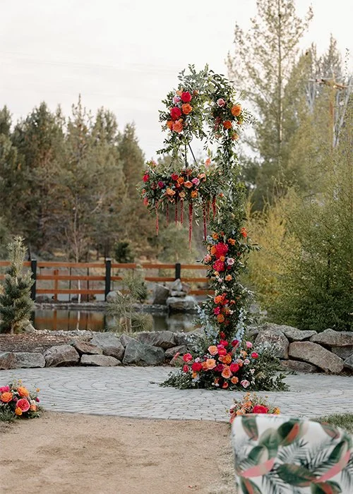 A floral wedding arch decorated with colorful flowers and greenery, set outdoors near a pond and surrounded by trees.