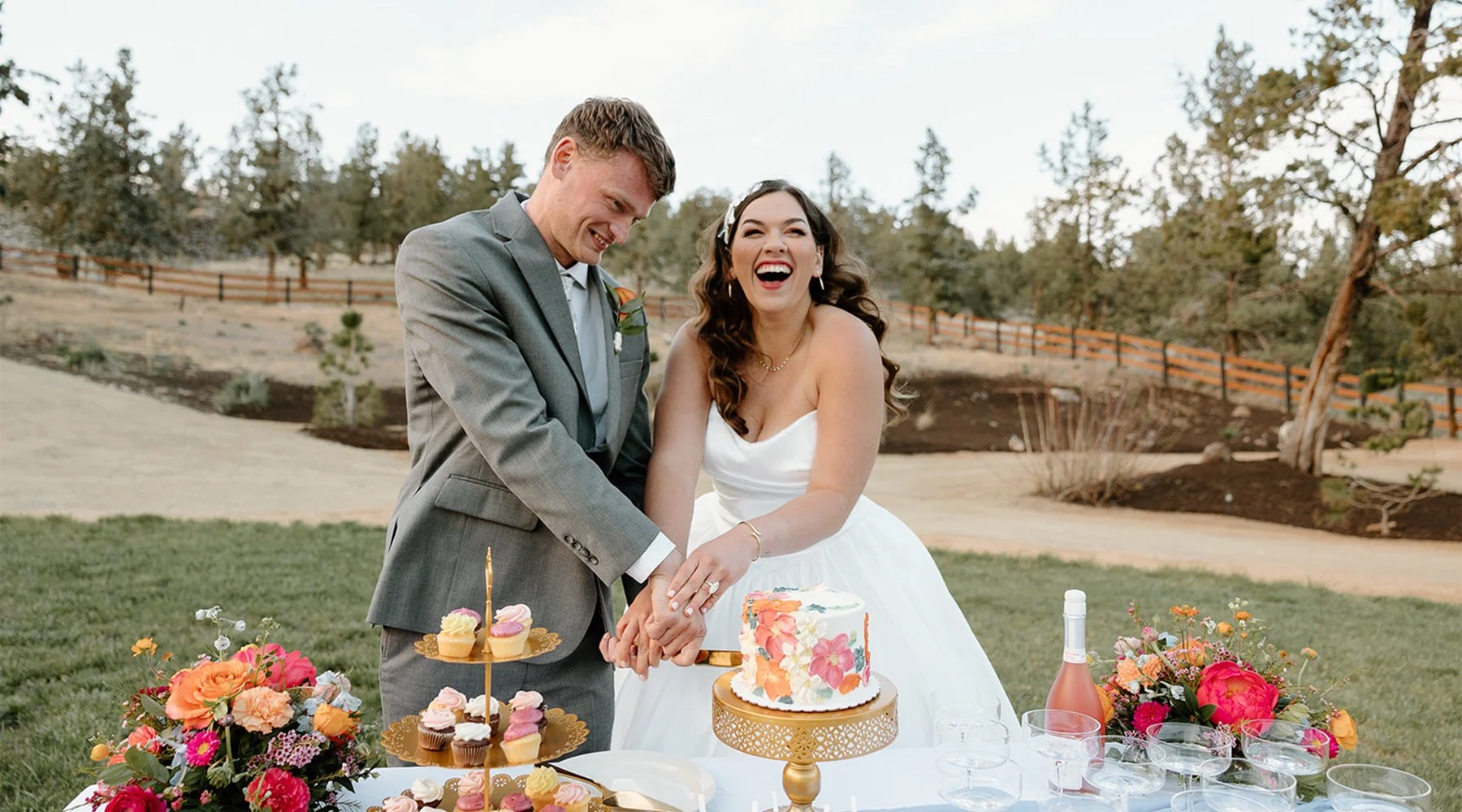 A bride and groom cutting a wedding cake outdoors, surrounded by flowers, cupcakes on a tiered tray, and drinks, with trees and a fence in the background.