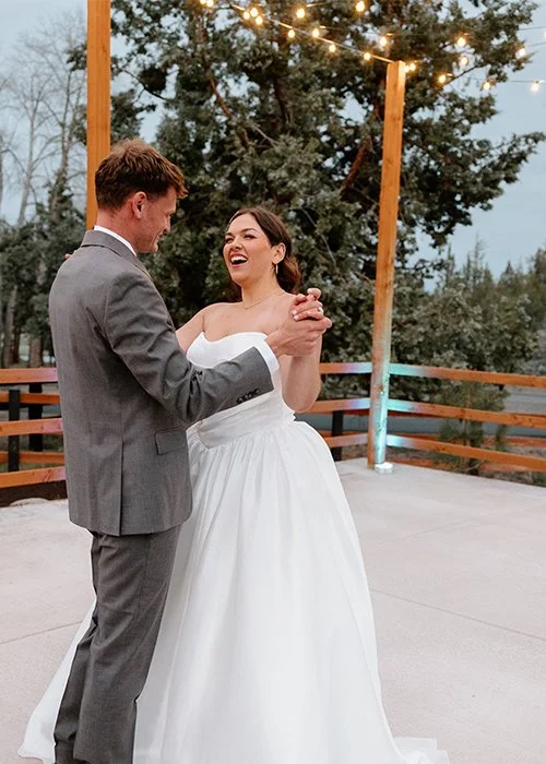 A bride and groom dancing outdoors during their wedding at dusk, with string lights and trees in the background.