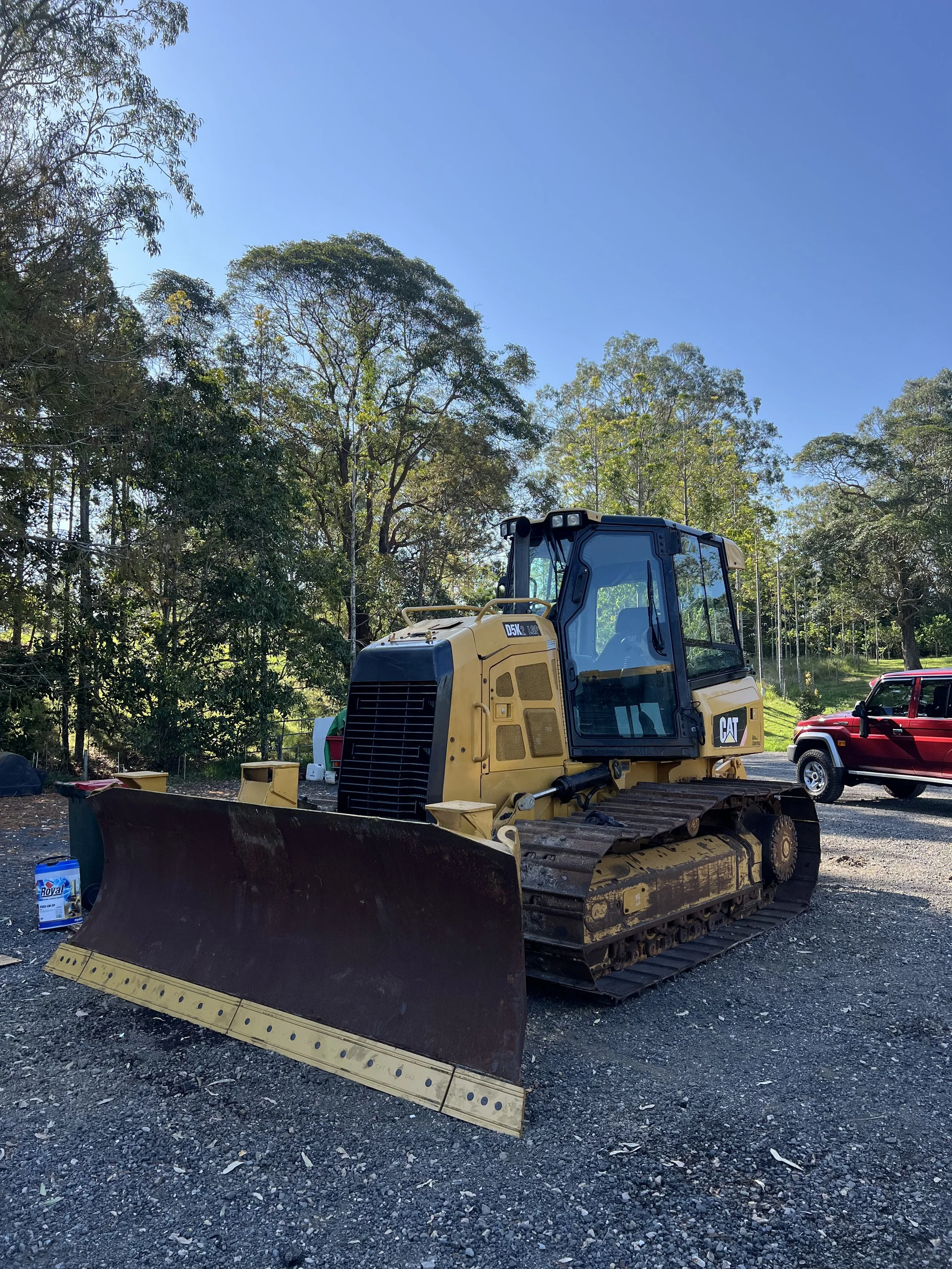A yellow Caterpillar bulldozer parked on rough gravel ground outdoors, with trees and a blue sky in the background.