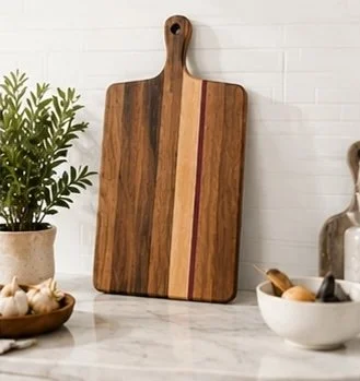 Wood cutting board with a handle leaning against a white tiled wall, surrounded by potted plant and bowls of garlic and eggs.