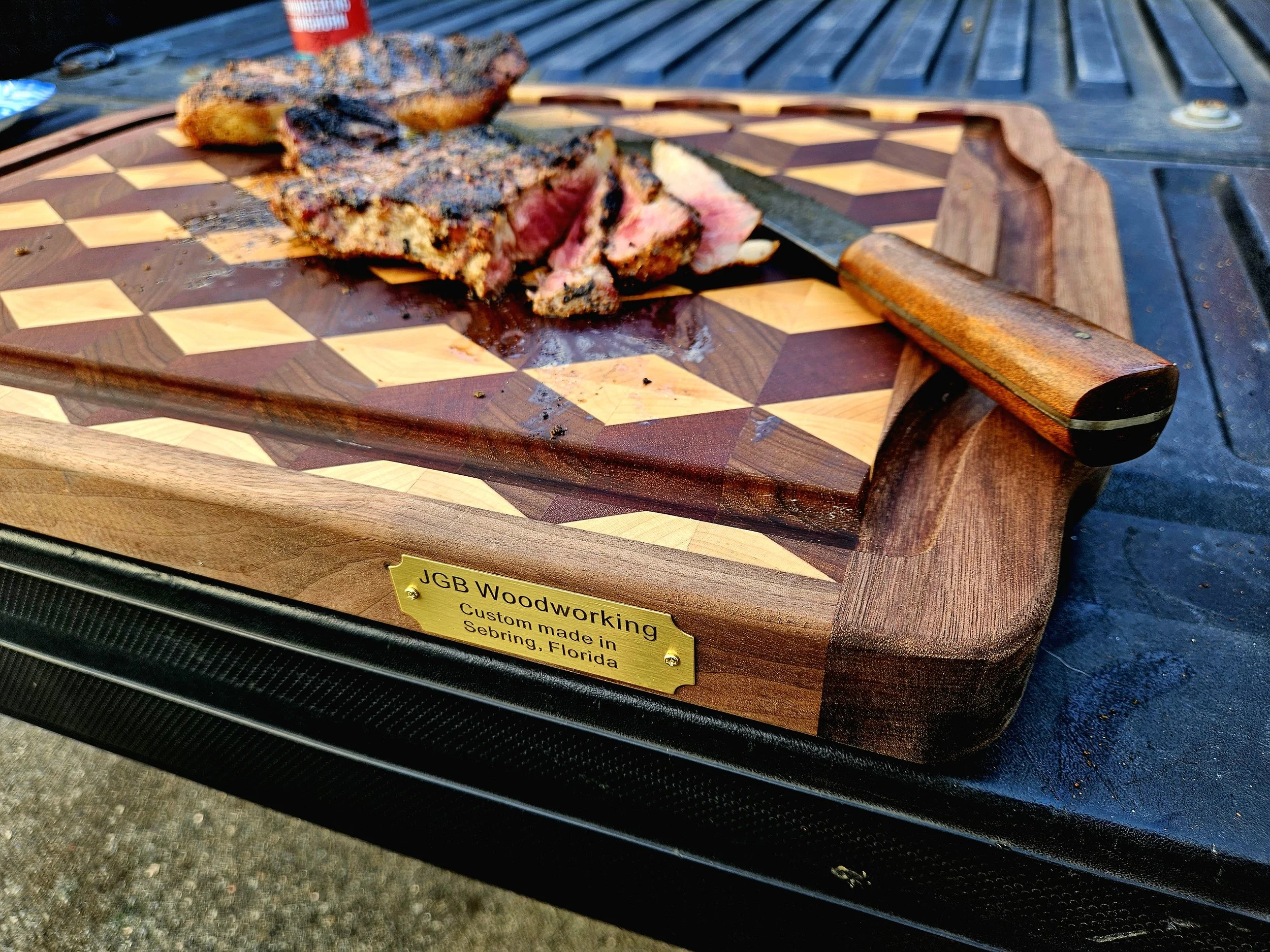 A partially eaten piece of grilled steak with a pink center on a decorative wooden cutting board with an intricate geometric pattern. A knife is placed beside the steak, and there is a yellow and black nameplate with the text "JGB Woodworking" and "Custom made in Sebring, Florida." The background shows a dark outdoor surface and a black table with a round metal object.