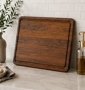 Empty wooden cutting board leaning against a white subway tile wall, with a glass soap dispenser and a plant in a vase nearby.