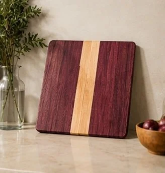 A wooden cutting board with a dark reddish-brown finish and a lighter vertical stripe in the middle, placed on a countertop with a potted plant and a wooden bowl with apples nearby.