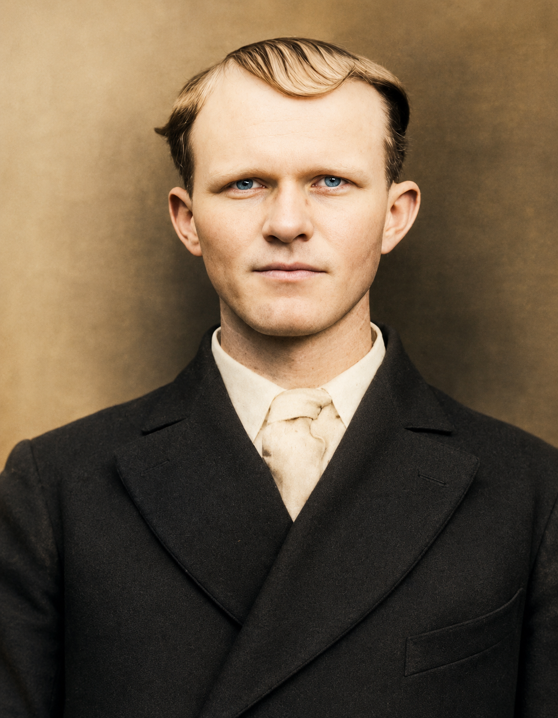 A young man with blond hair and blue eyes wearing a black suit with a white shirt and a light-colored tie, standing against a plain beige background.