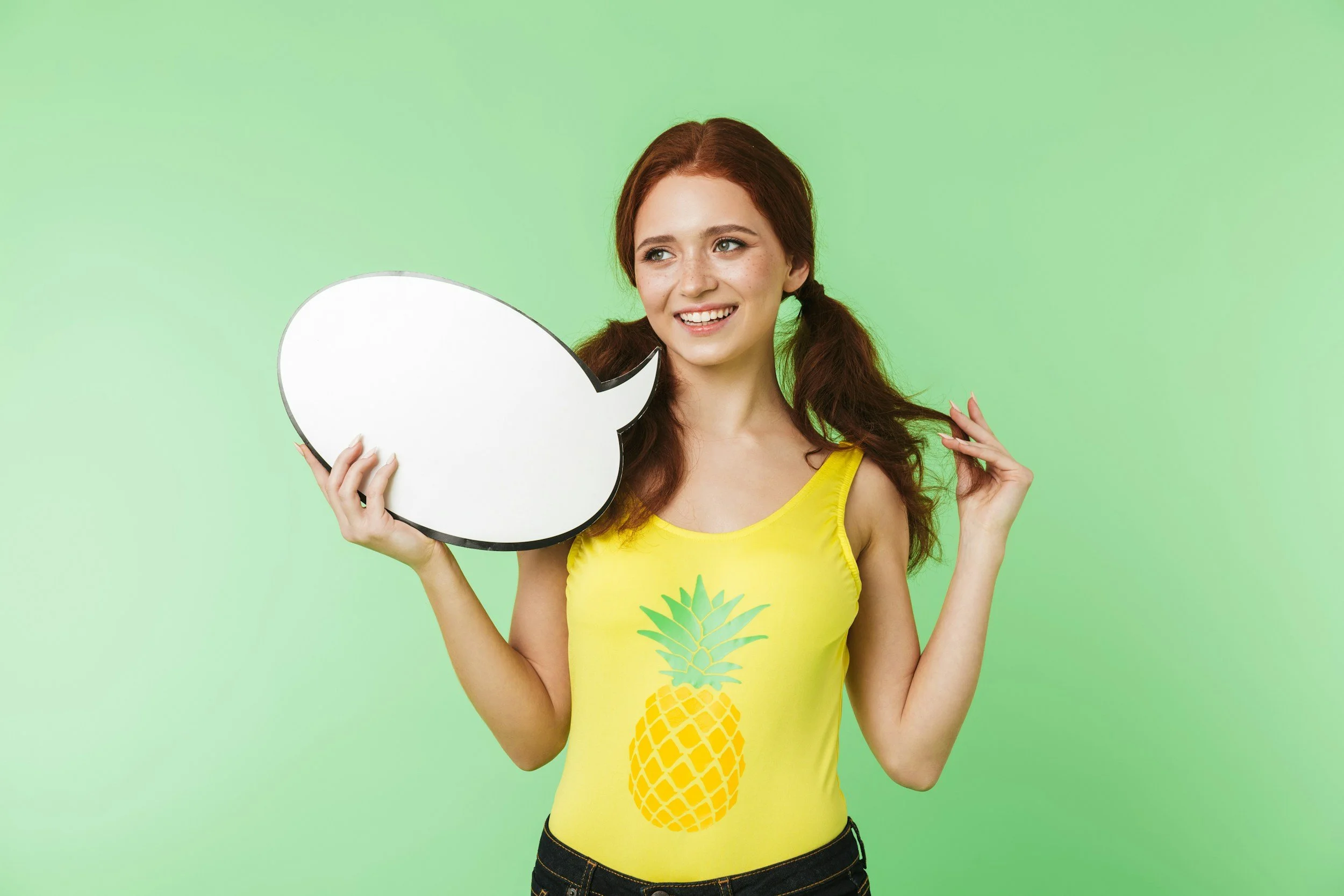 Junge Frau mit roten Haaren trägt gelbes Shirt mit Ananas-Design und hält eine leere Sprechblase, vor grünem Hintergrund.