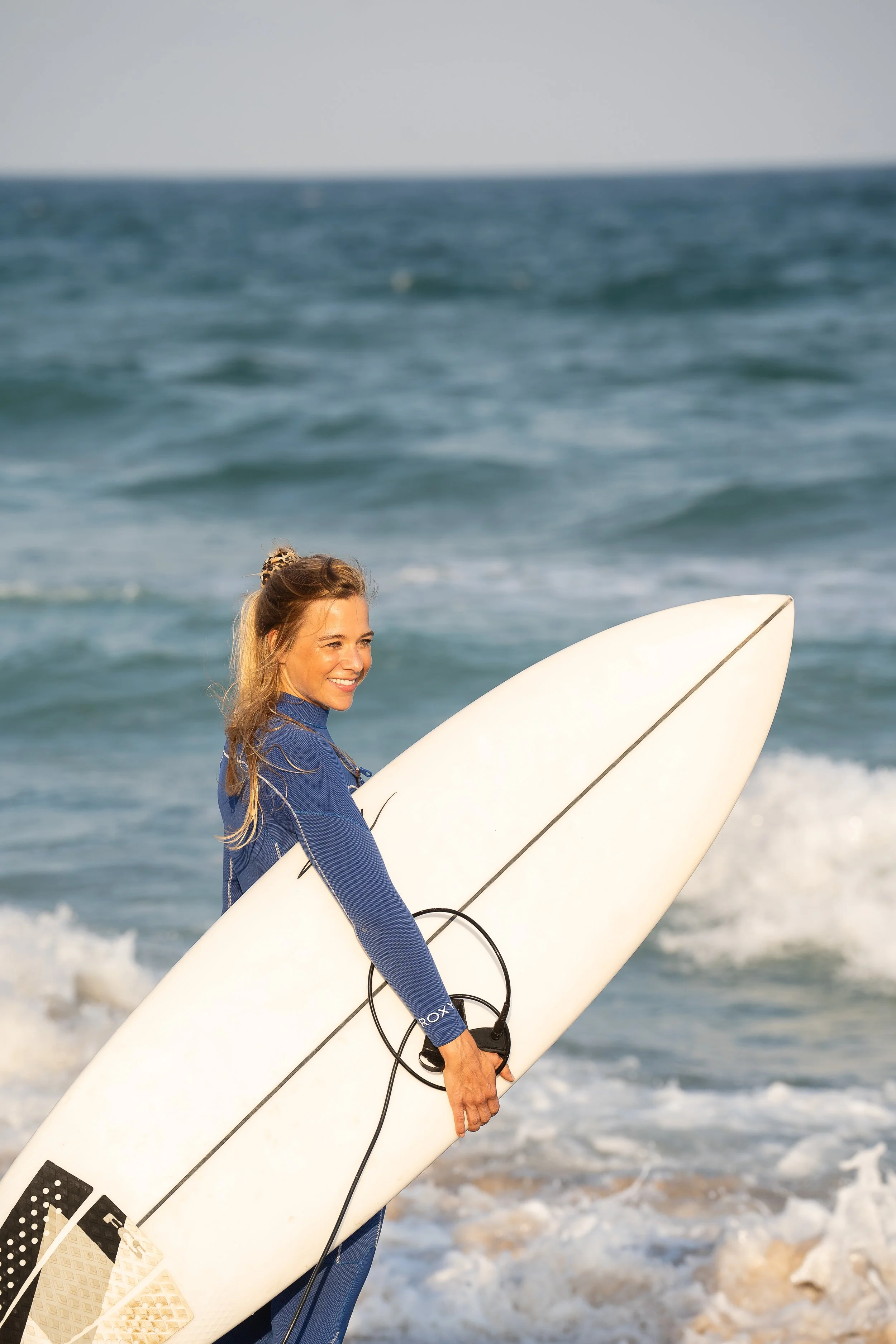 Eine lachende Frau mit langen blonden Haaren hält ein Surfbrett an einem Strand, das Meer im Hintergrund, sonniges Wetter.