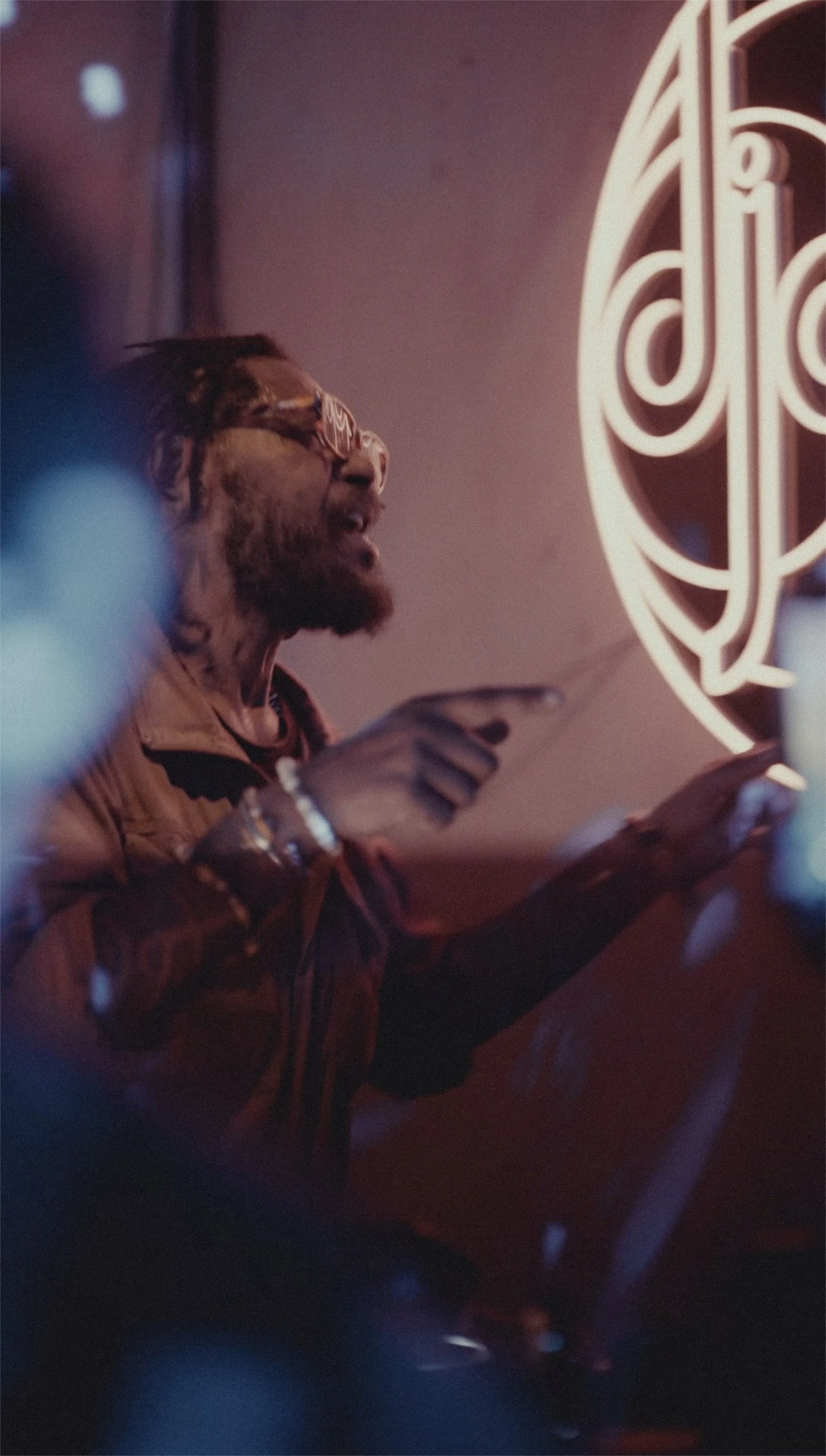 Homme avec des dreadlocks portant des lunettes de soleil, utilisant un téléphone portable, dans un lieu avec un éclairage rouge et une enseigne lumineuse en arrière-plan.