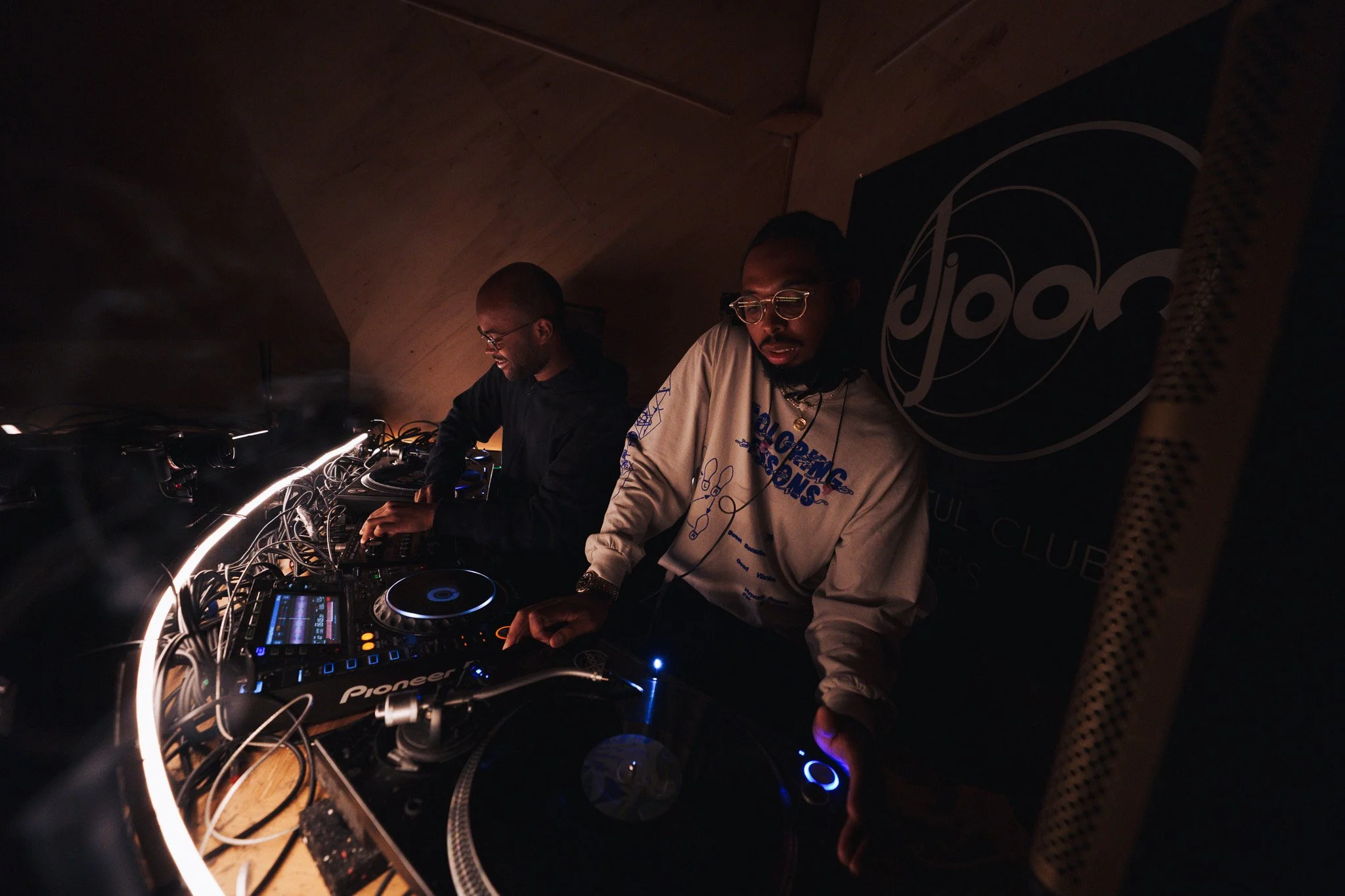 Deux DJ en train de mixer sur une platine DJ dans un espace sombre, avec un décor et un logo ' groove' en arrière-plan.