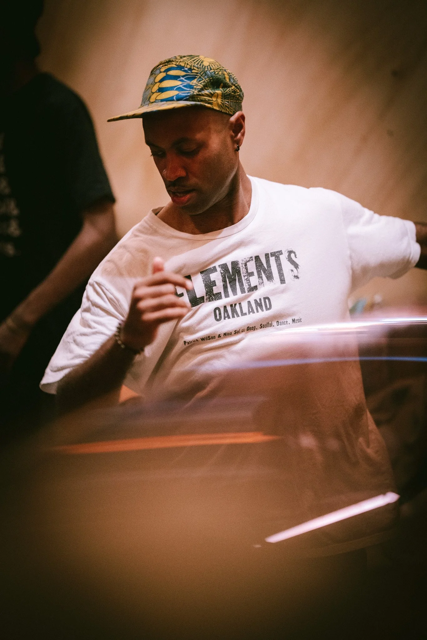 Un homme portant un chapeau coloré et un t-shirt blanc avec des inscriptions noir, semble danser ou faire du mouvement dans un endroit sombre ou tamisé.