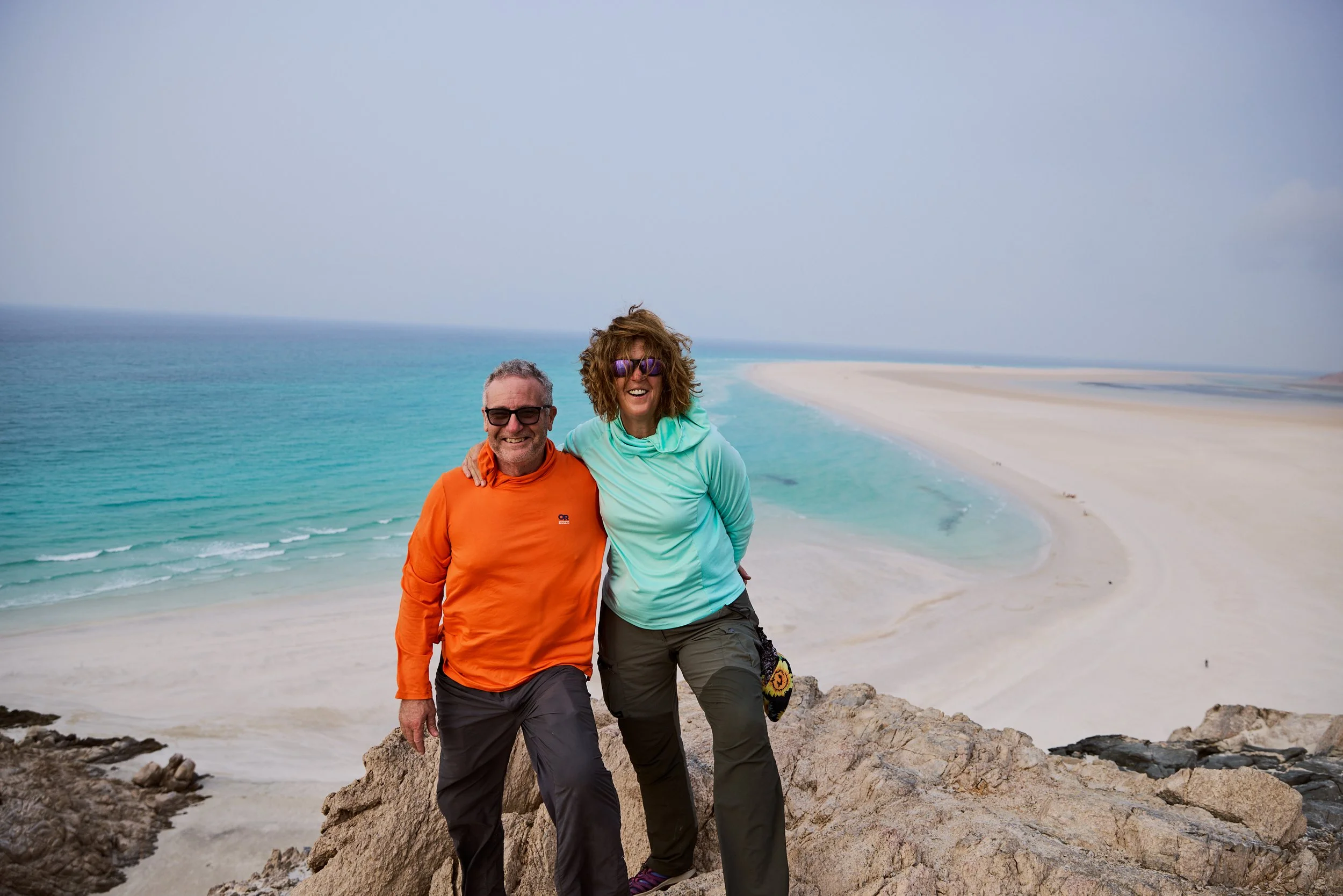 A smiling older man and woman standing on rocks at the beach, with the ocean and a sandy shoreline in the background. They are dressed in outdoor clothing suitable for cool weather.