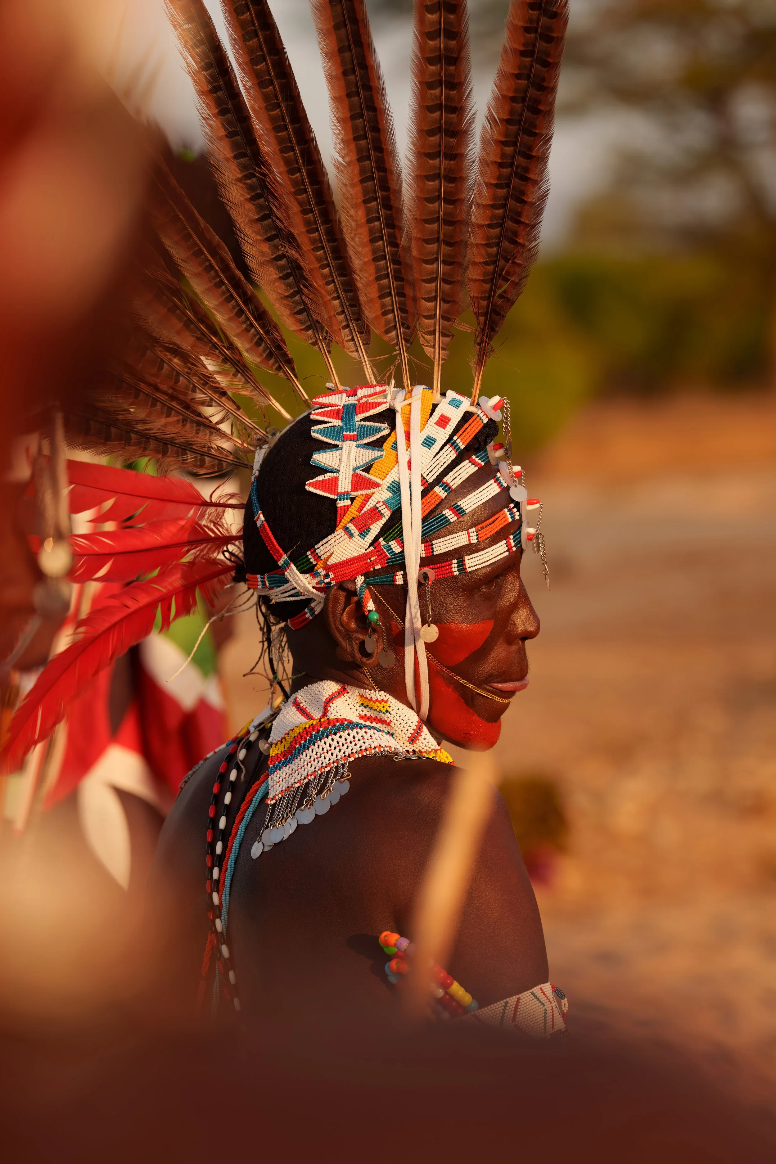 A Samburu Warrior in wearing traditional colorful bead jewelry and a headdress with brown feathers. The person has facial paint around their nose and wears multiple necklaces and earrings. Northern Kenya, Kalepo camp