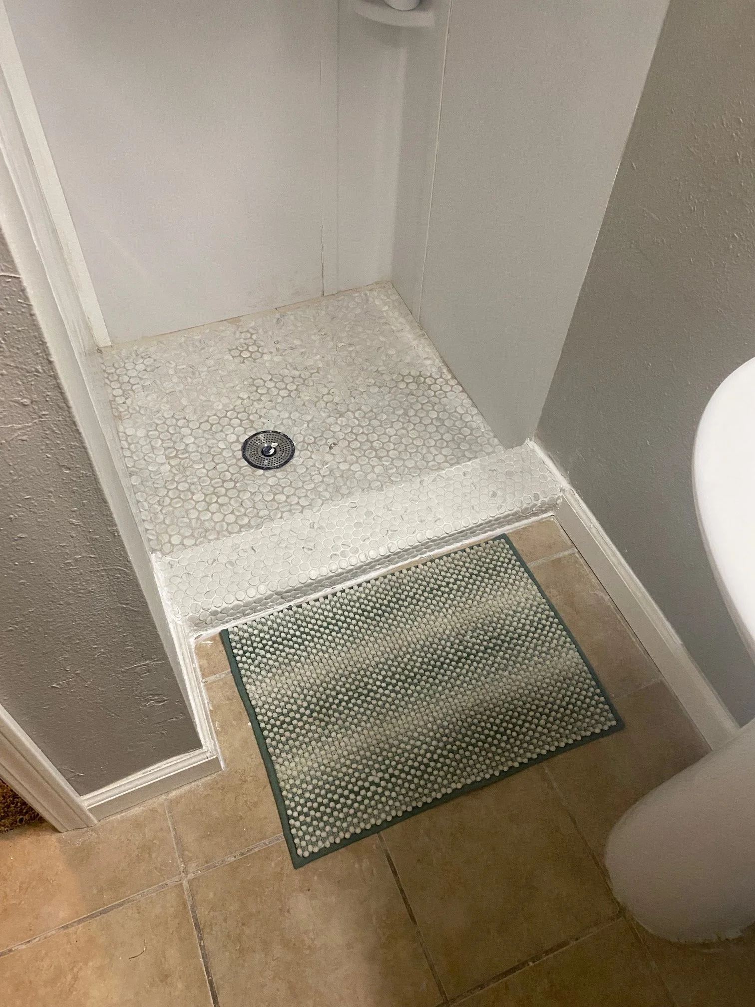 Empty shower with white tiled walls, a round metal drain on the floor, and a beige tile floor with a small rectangular green and beige bath mat nearby. Part of a white toilet is visible on the right side.