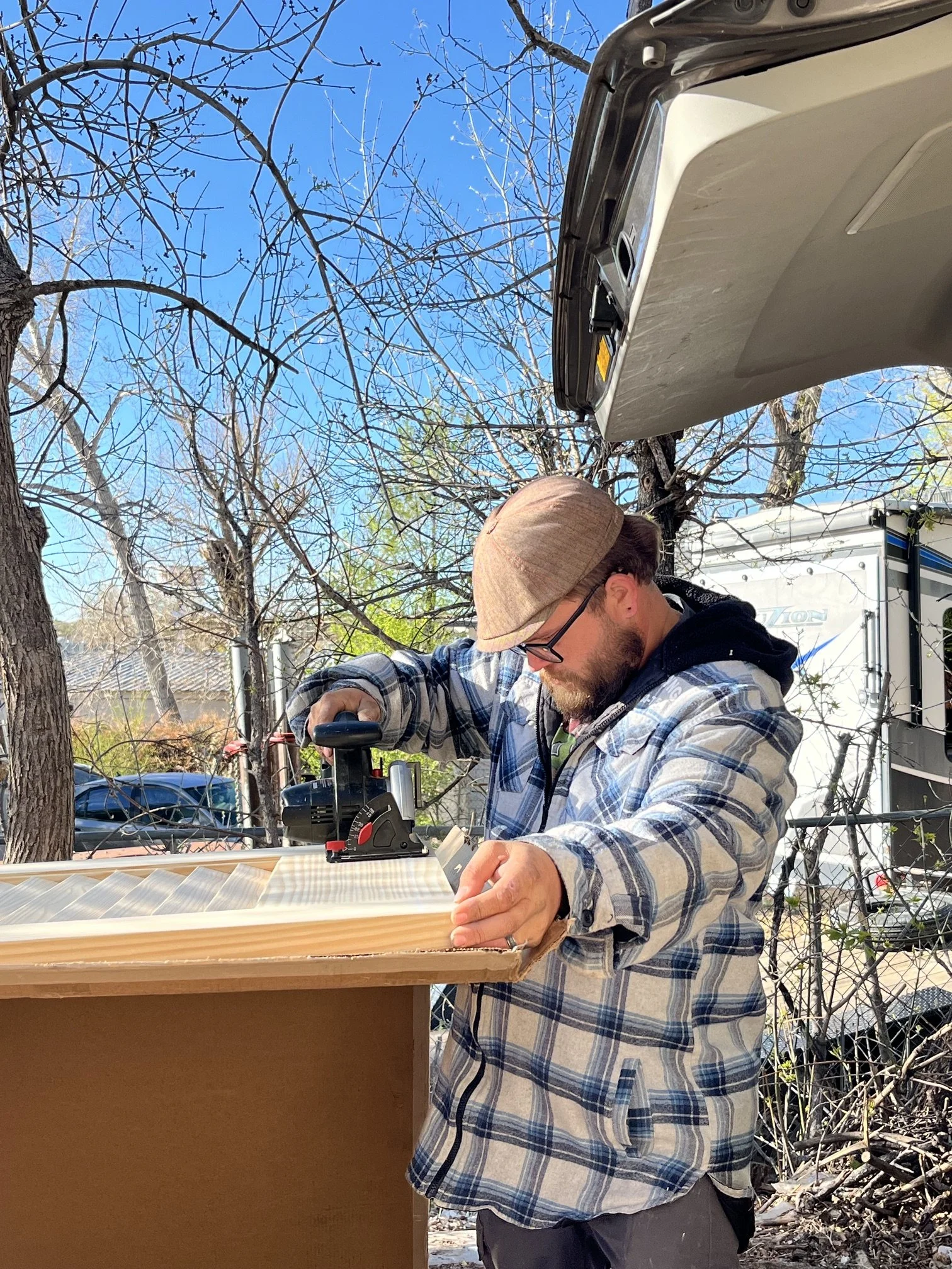 A man with a beard, glasses, and a tan hat working on a wooden frame outdoors during daytime. He is wearing a plaid shirt and is focused on assembling or adjusting the structure. There are trees with leafless branches in the background and a blue sky.