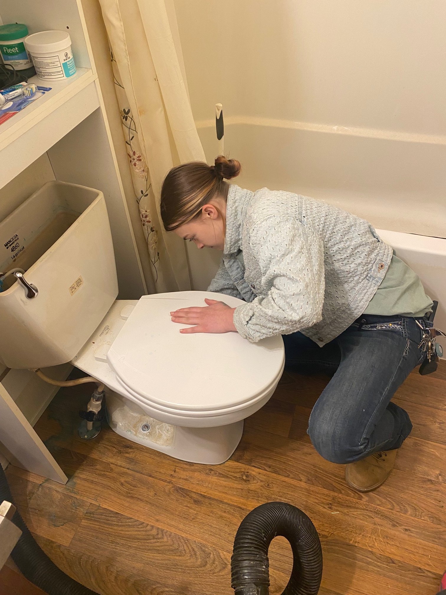 A person is kneeling in front of a toilet in a bathroom, leaning forward with their hands on the toilet seat, wearing a gray jacket, glasses, and jeans, with a set of keys hanging from their pocket.