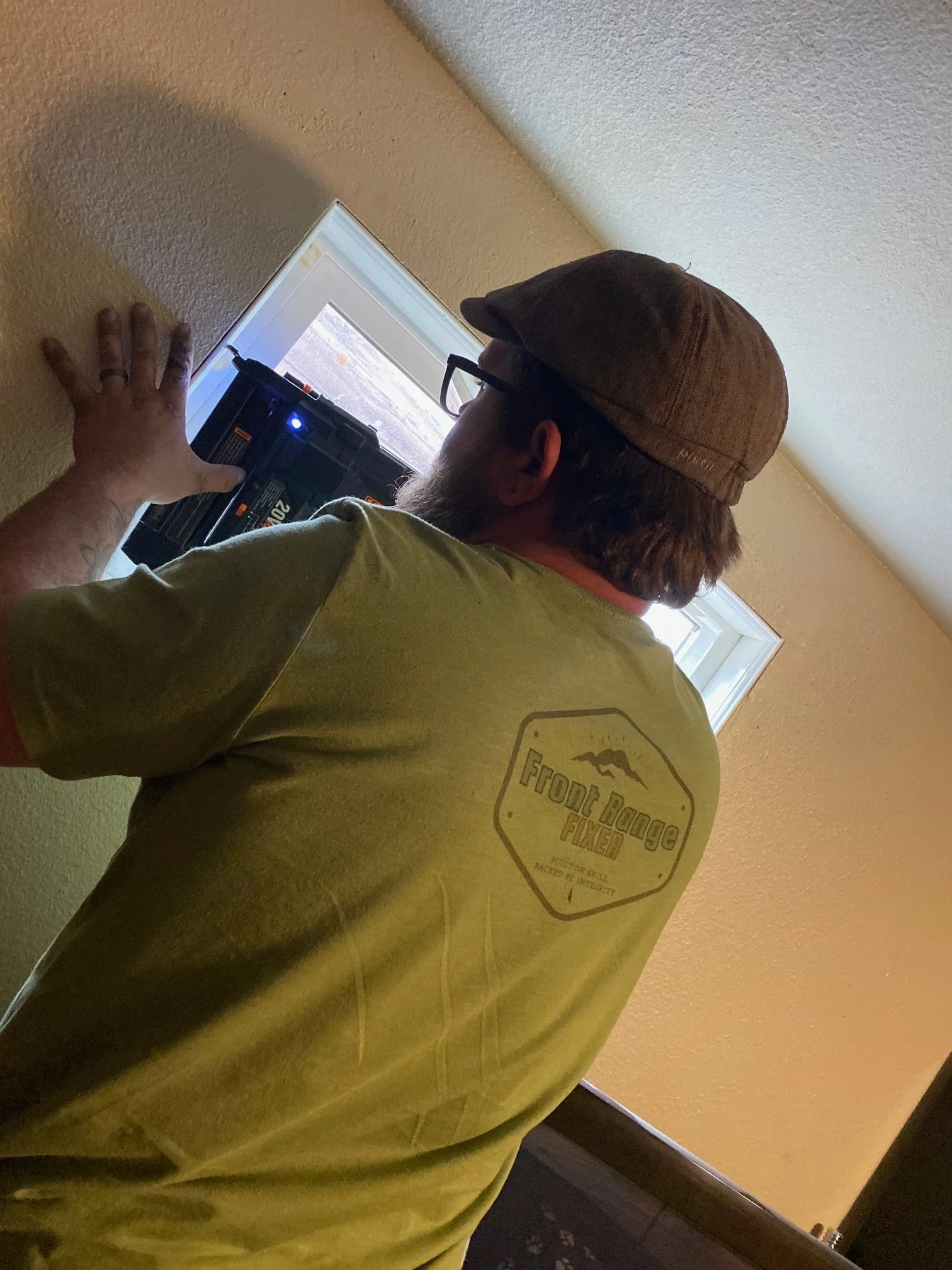 Side view of a man wearing glasses, a brown cap, and a yellow T-shirt with a logo that reads 'Front Range Fixer,' standing indoors near a window with beige walls.