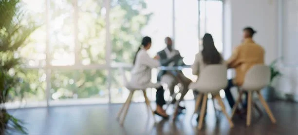 A group of five diverse people sitting around a table in a modern meeting room with large windows and greenery outside.