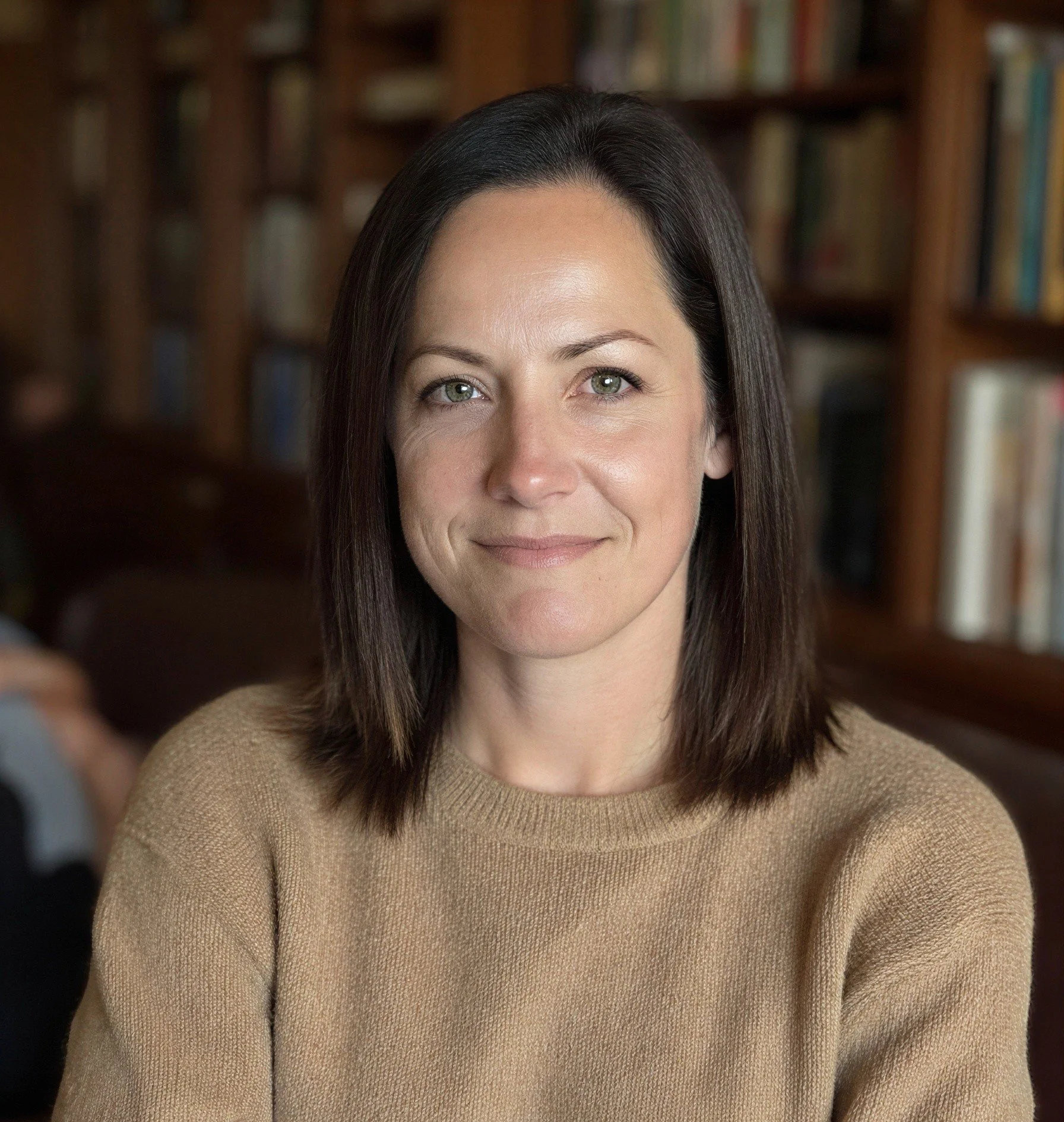 Close-up of a woman with shoulder-length brown hair, green eyes, and light skin, smiling softly, wearing a beige sweater, with a background of bookshelves filled with books.
