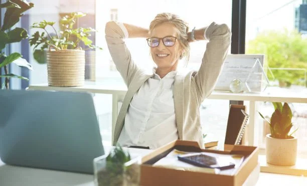 A woman with glasses smiling and relaxing at a bright office desk with plants, a laptop, and office supplies.