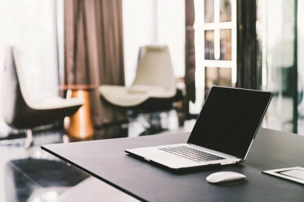 Open laptop, mouse, and tablet on a conference table in a modern office with chairs and windows in the background.