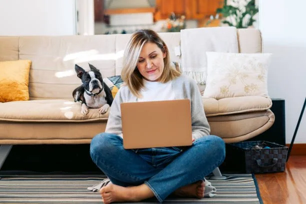 A woman sitting on the floor with a laptop on her lap, with a Boston Terrier dog sitting on a sofa behind her in a living room.