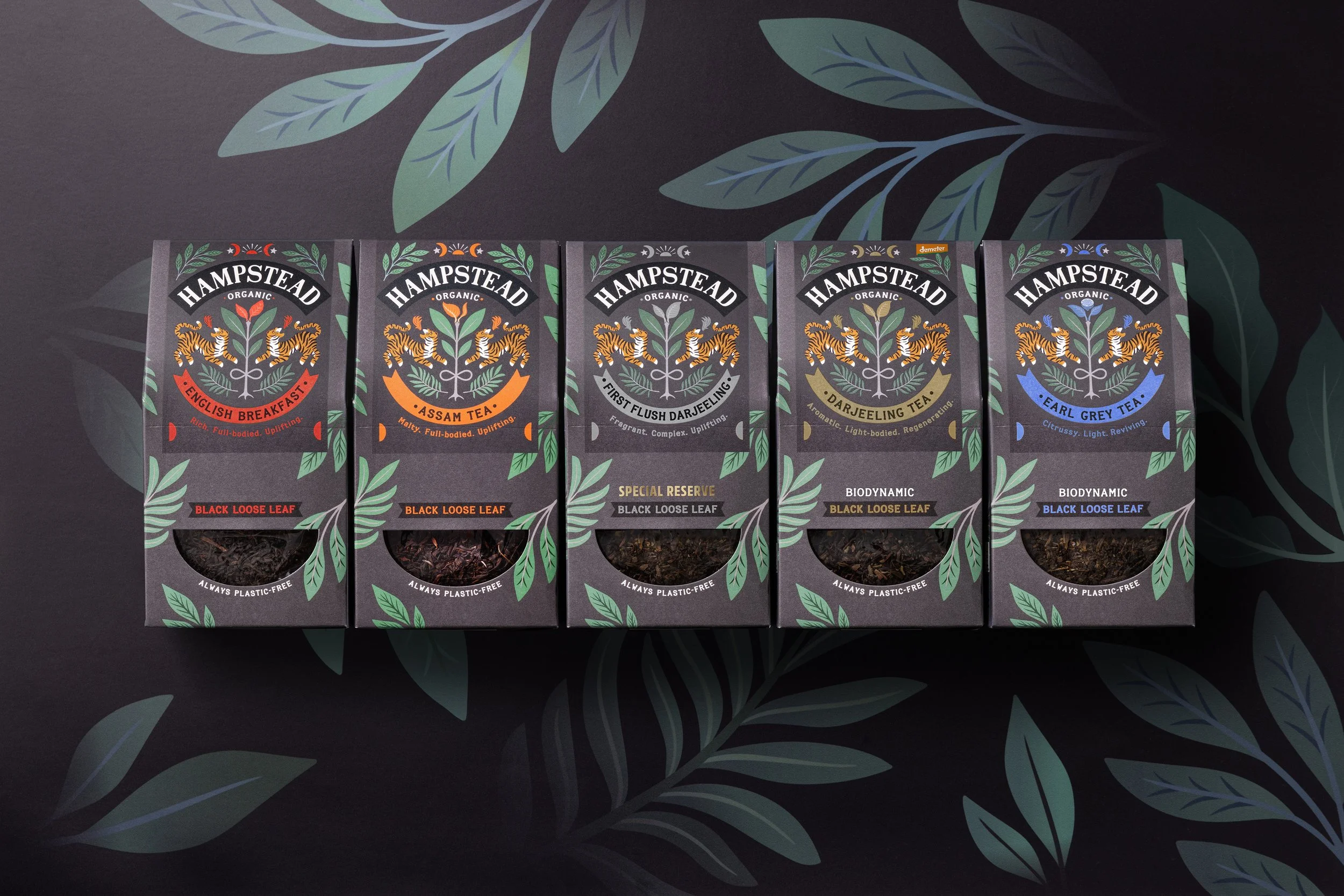 A row of five black tea boxes from Hampstead Organic with different flavor labels, set on a dark surface with large leaf patterns.