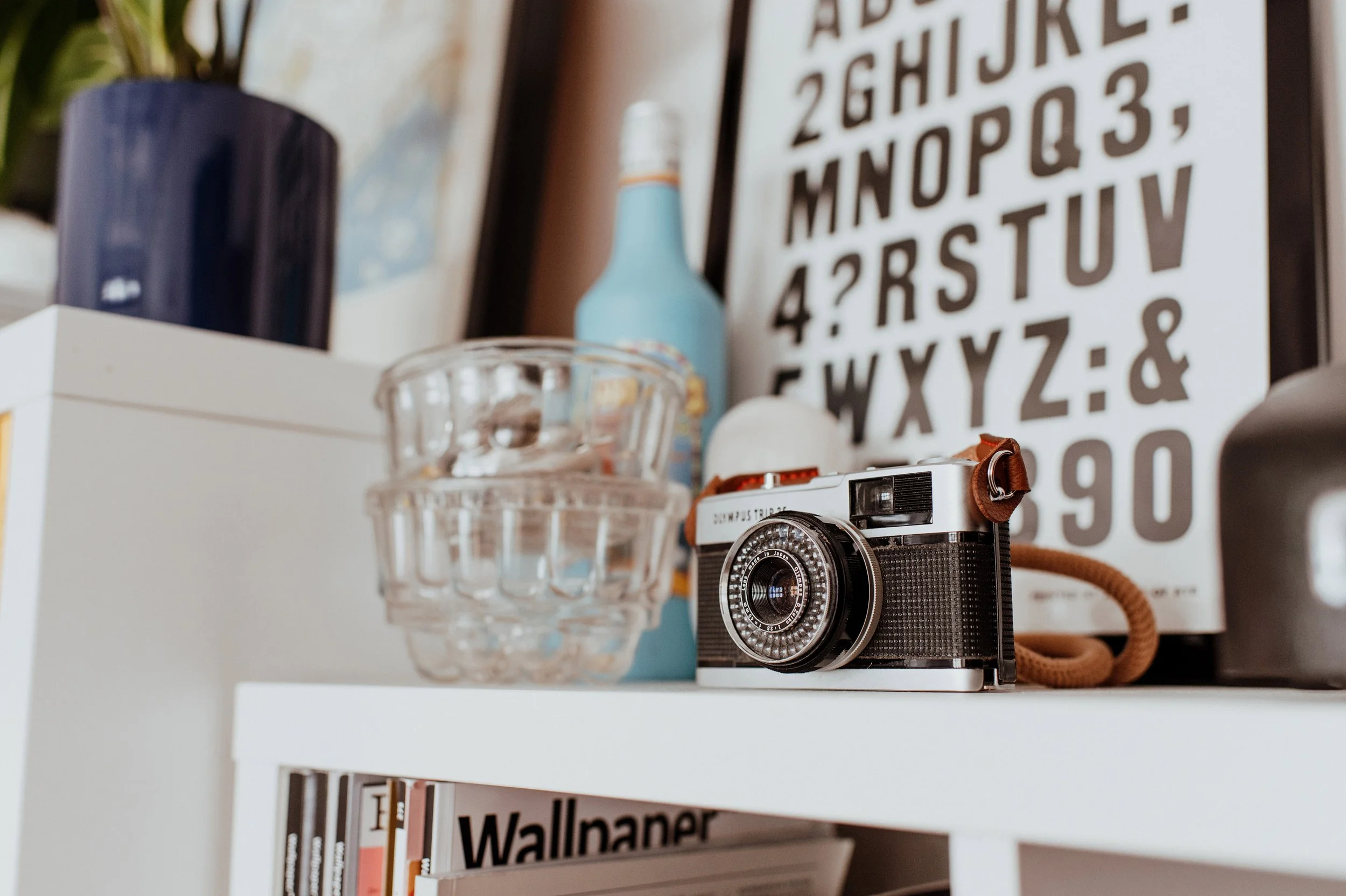 Shelf with various objects including a vintage camera, a glass container, a blue bottle, a framed print with alphabet letters, and wall-mounted wallpaper books.