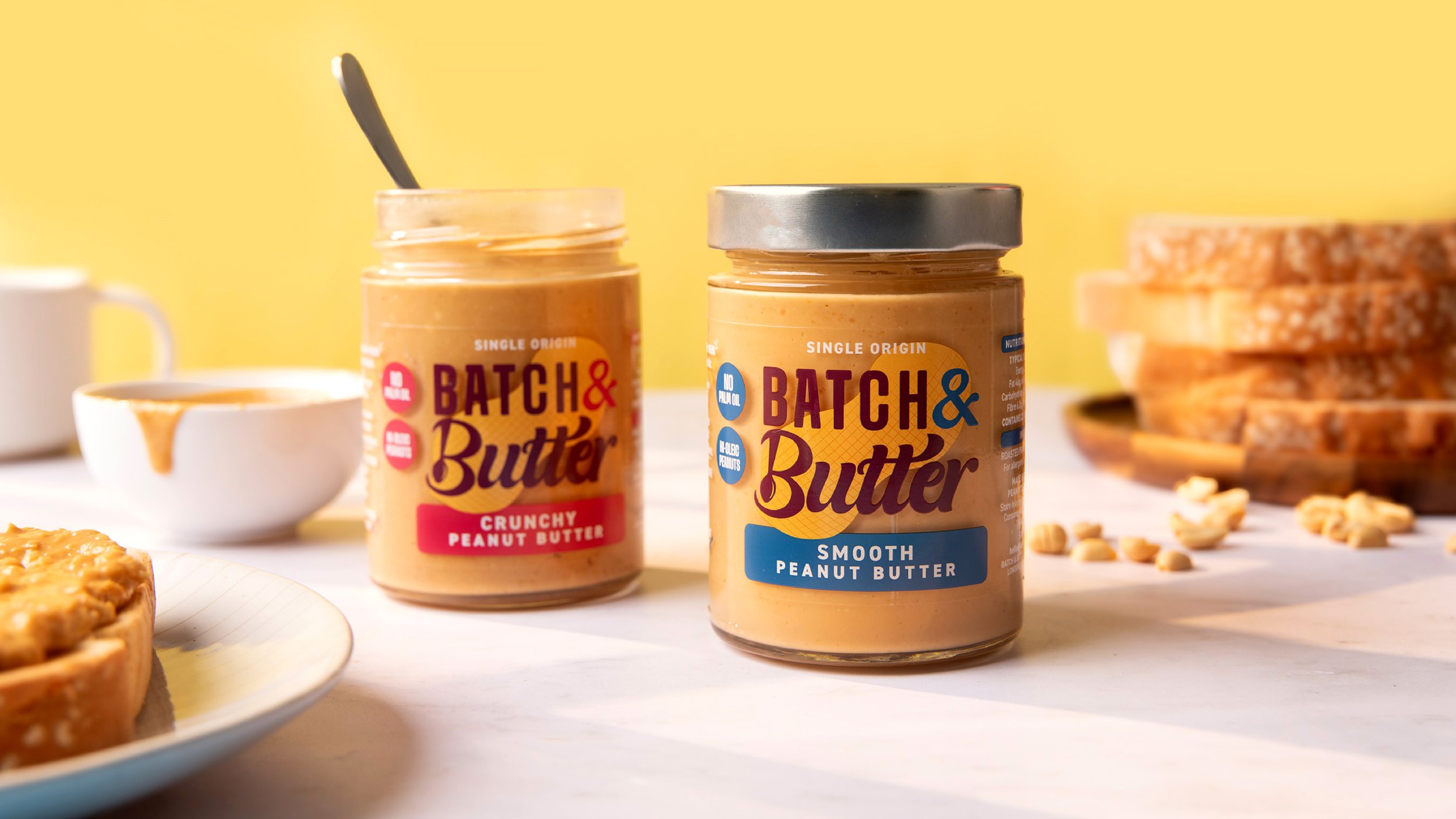 Two jars of Batch & Butter peanut butter, one labeled crunchy and one smooth, with a bowl of peanut butter and a plate of granola bars in the background.