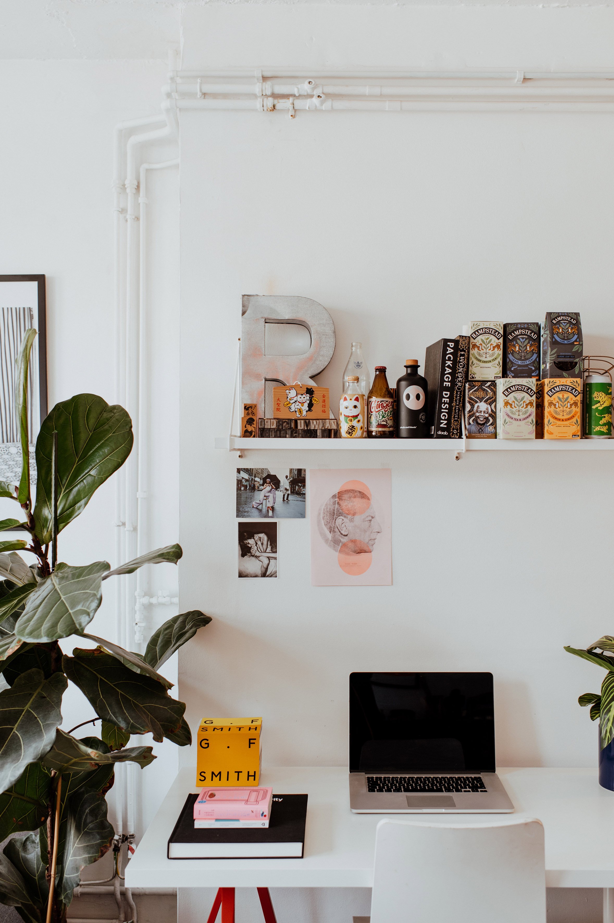 A clean, modern workspace with a white desk holding a MacBook, a black notebook, a pink are, and a yellow box with branding. Behind the desk is a white wall with a shelf displaying various books, boxes, and decorative items, including a large metal l