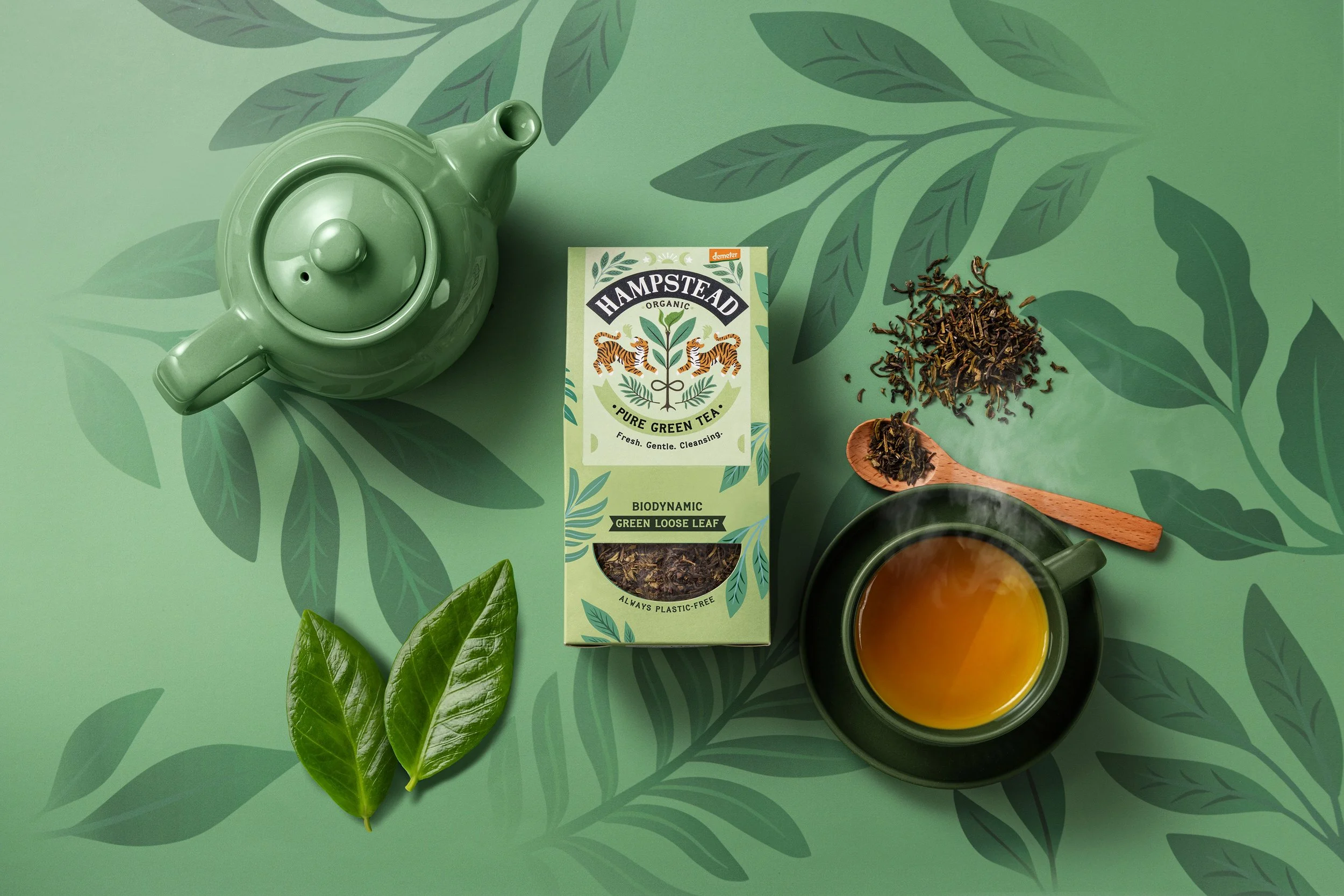 A green teapot, a box of Hampstead organic green loose leaf tea, loose tea leaves, a cup of tea, and green tea leaves on a green leafy patterned surface.