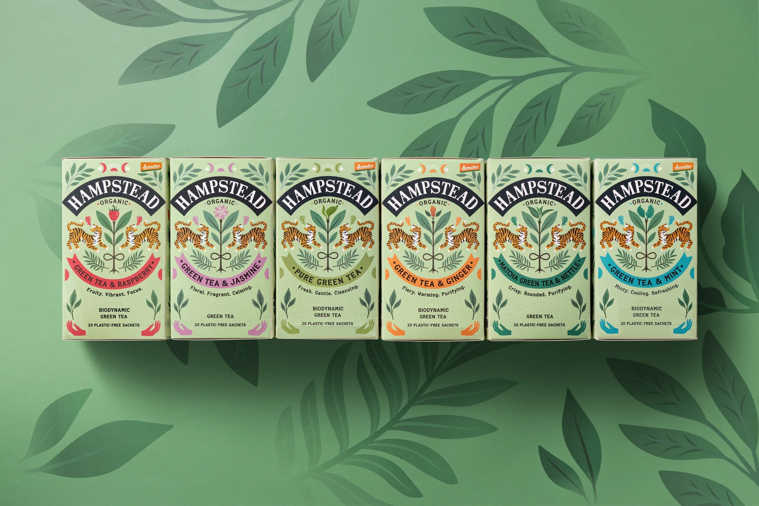 A row of six boxes of Hampstead organic green tea with floral and tiger illustrations, placed on a green surface with leaf patterns in the background.