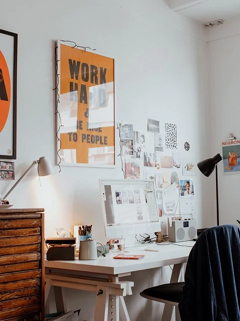 A tidy home workspace with a white desk, computer monitor, desk lamp, and various office supplies, against a white wall decorated with posters and photos.