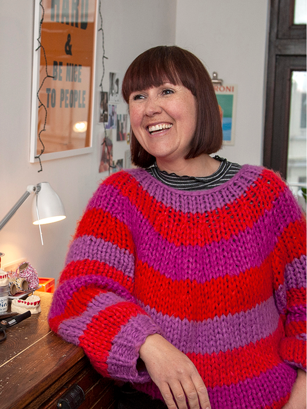 Woman with short brown hair smiling while wearing a chunky, colorful striped sweater in red, purple, and pink, sitting in a cozy room with a desk lamp and decorative items on a wooden table.