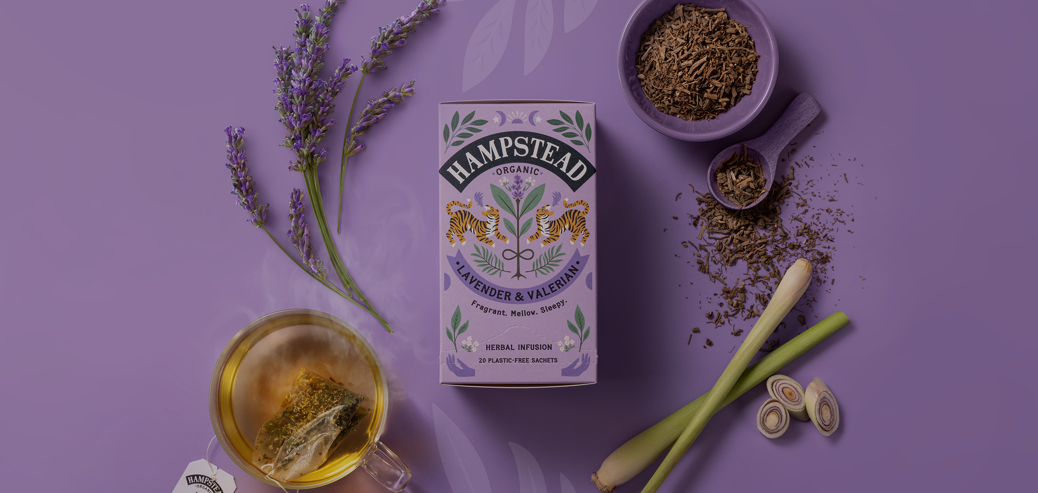 A box of Hampstead Organic Lavender and Valerian herbal infusion tea surrounded by lavender sprigs, a cup of tea with a tea bag, dried herbs in a purple bowl, a scoop with dried herbs, and sliced leek on a purple surface.