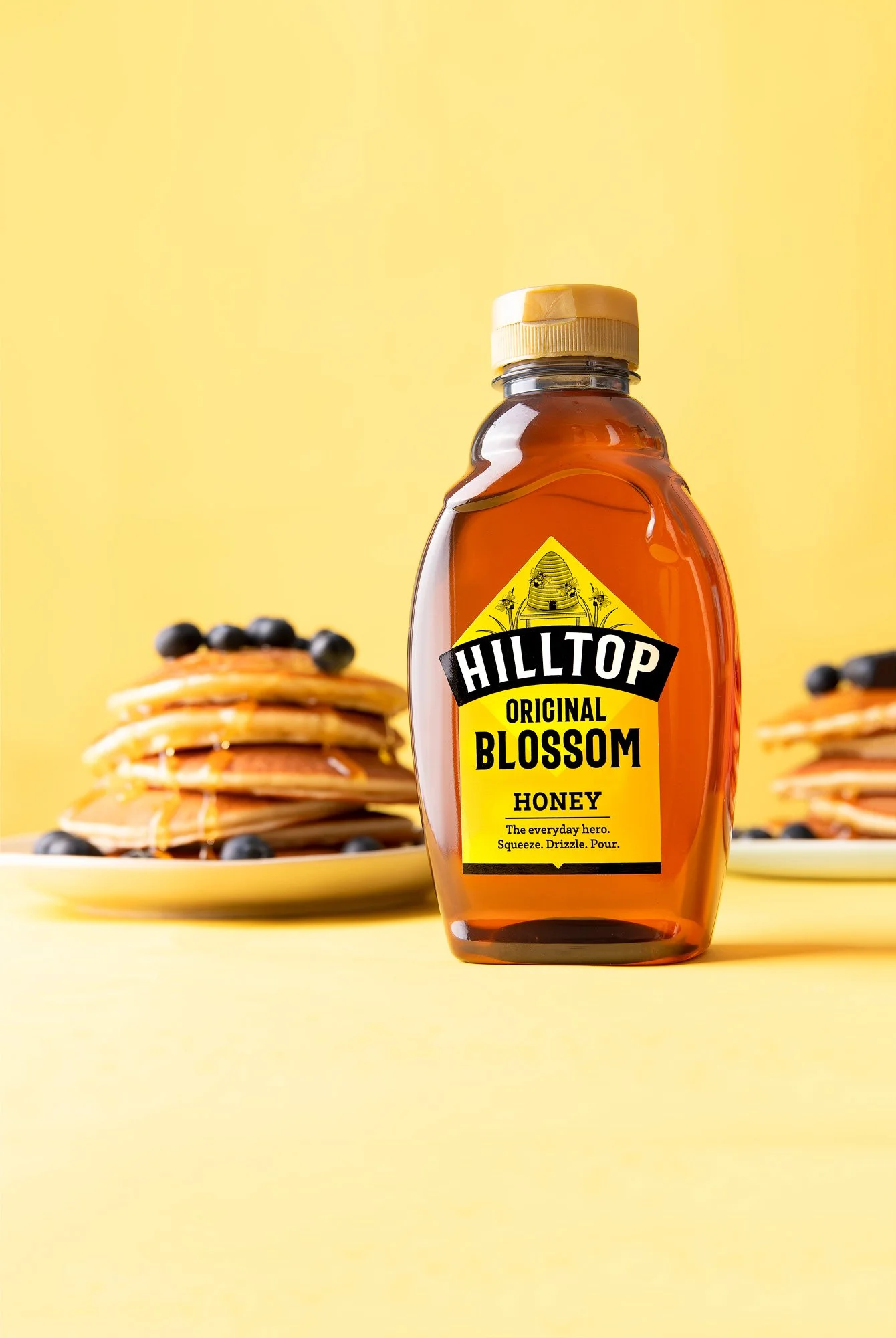 A bottle of Hilltop Original Blossom honey with a yellow label in front of a stack of pancakes topped with blueberries on a yellow background.