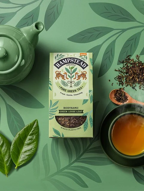 A box of Hampstead organic green loose leaf tea, a teacup of brewed tea, a green teapot, green leaves, and loose tea leaves on a green leafy background.