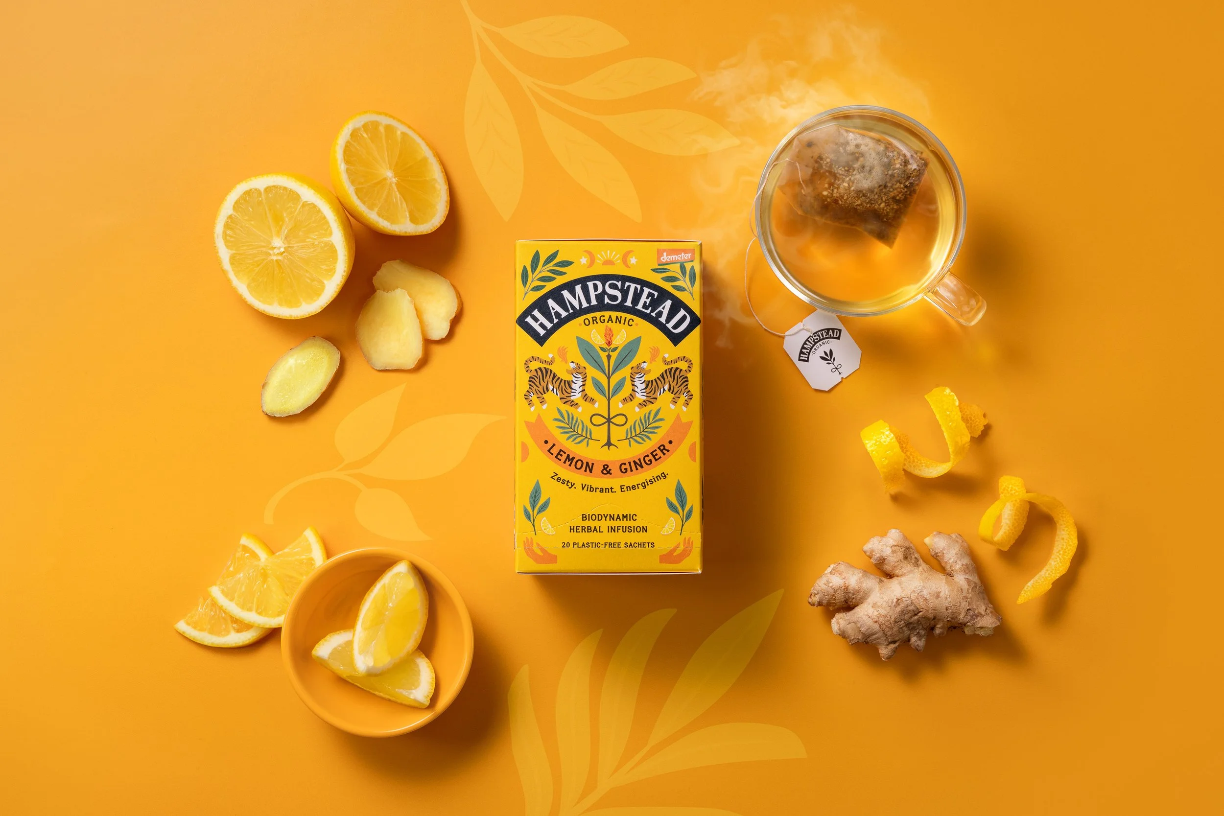A yellow box of Hampstead Lemon & Ginger herbal infusion tea surrounded by fresh lemon slices and ginger root on a yellow background.