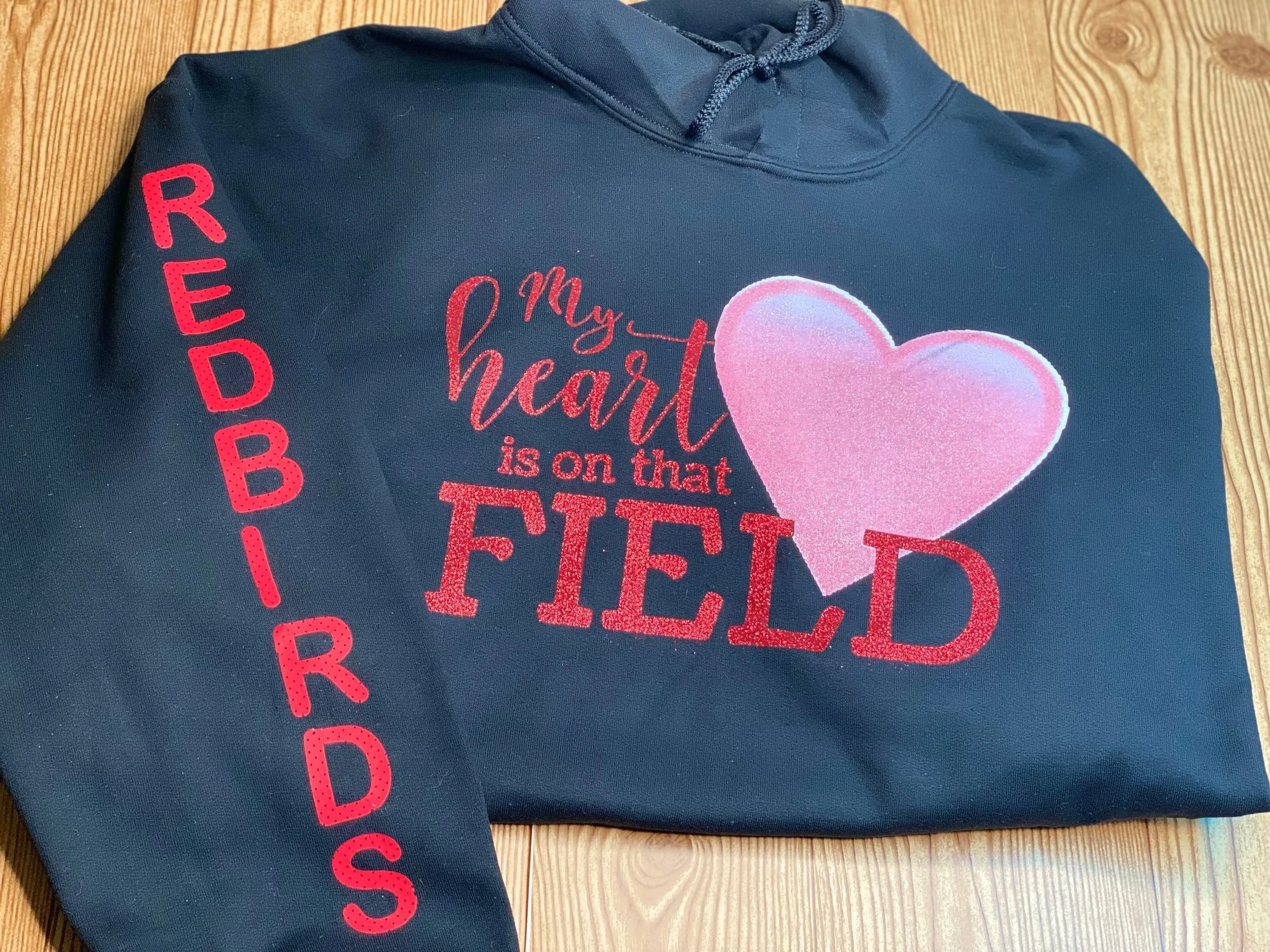 A navy blue hoodie with red and pink embroidered text and a pink heart. The text says 'My heart is on that TIED'. The left sleeve has the word 'REDDI' vertically in red. The hoodie is on a wooden surface.