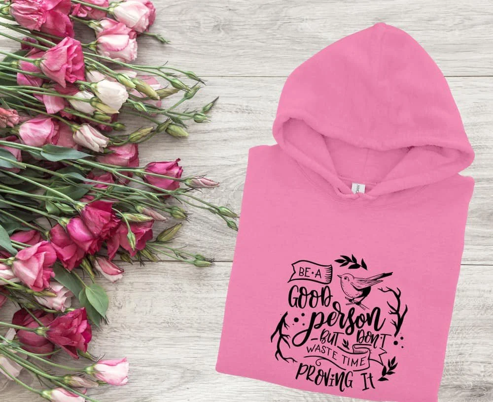 Pink hoodie with a motivational quote and a bird illustration next to a bouquet of pink and white flowers on a light gray wooden surface.