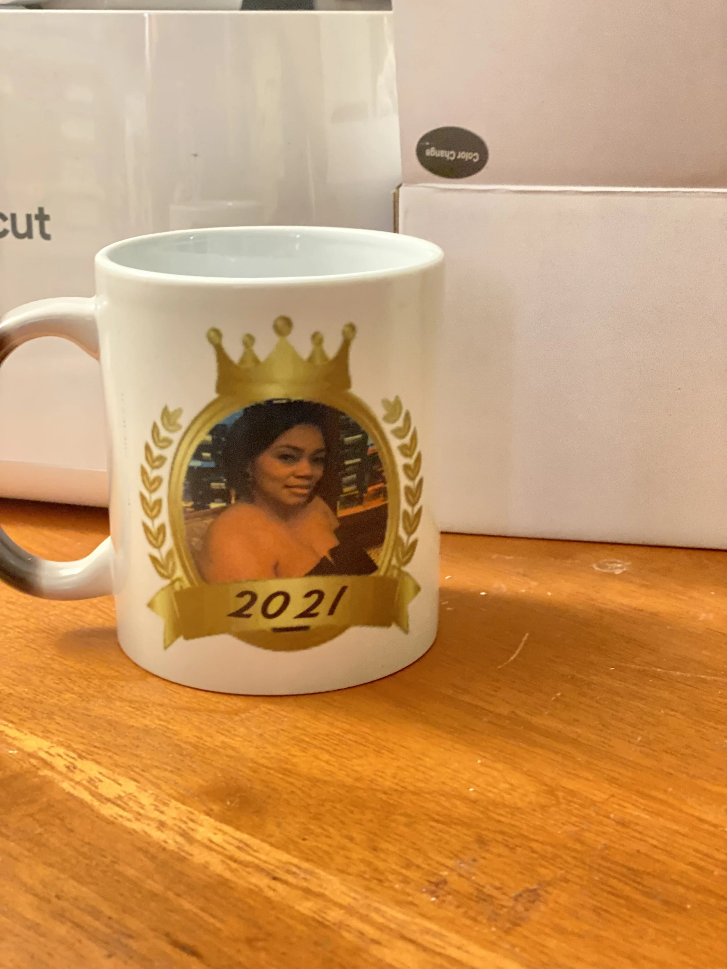 A white ceramic mug featuring a woman with dark hair, wearing an orange shirt, framed by a gold crown and laurel design, with the year '2021' on a gold banner. In the background, there are boxes, one labeled 'Color Change' and another partially visib