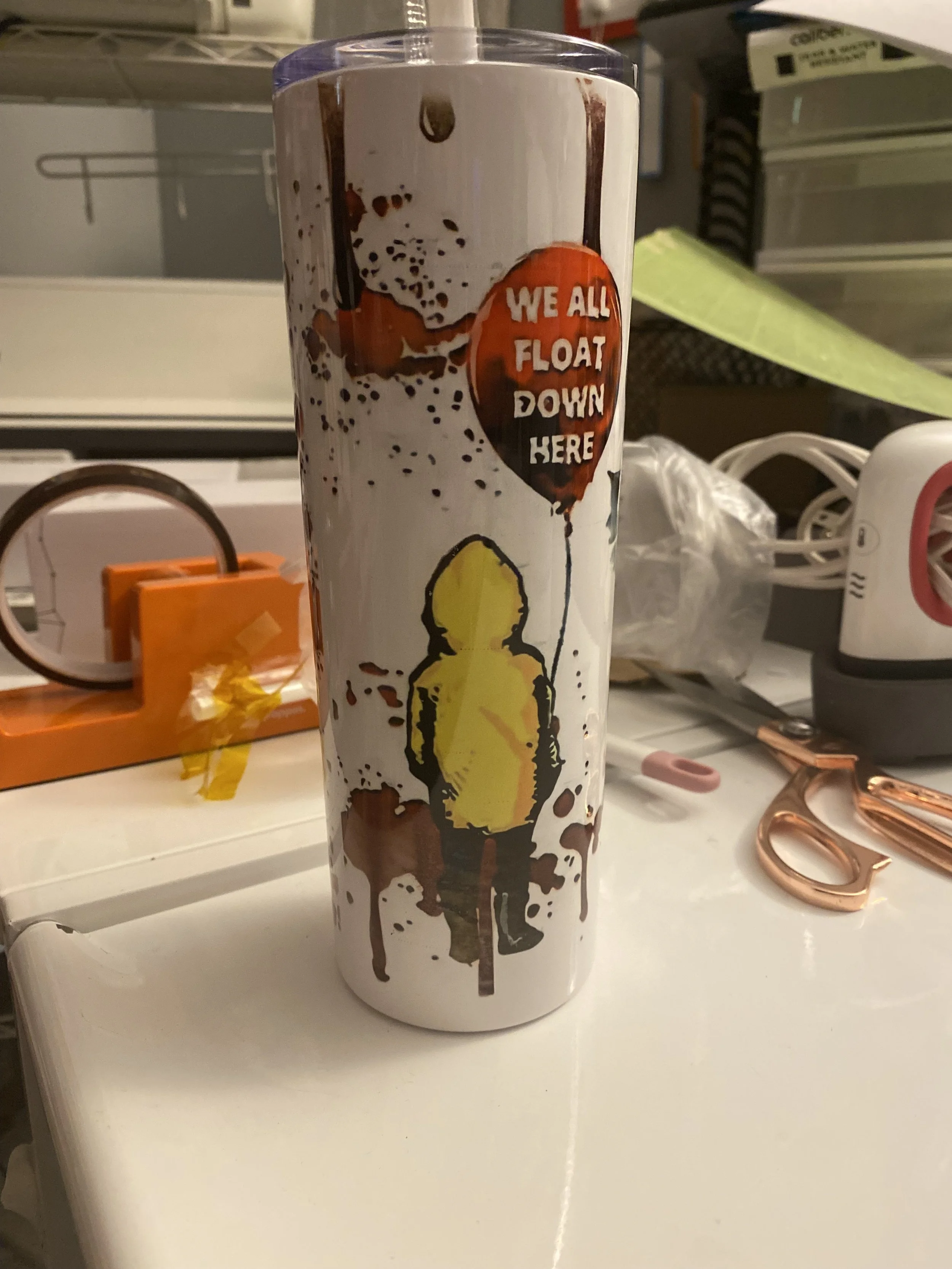 A tumbler with a graphic of a person on it, holding a balloon-shaped sign that reads 'We All Float Down Here', placed on a white surface with various objects and clutter in the background.