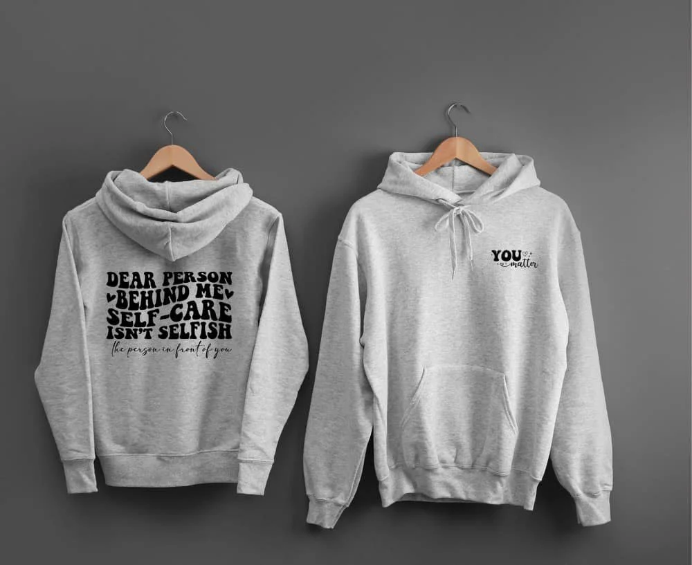 Two gray hoodies hanging on wooden hangers against a gray wall. The hoodie on the left has text on the back that reads, "Dear person behind me, self-care isn't selfish. The person in front of you." The hoodie on the right has text on the front that says, "You" with small hearts and stars around it, and the word "smaller" underneath.